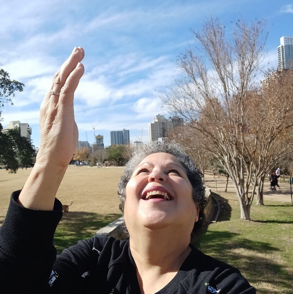 Foto de Carmen Minerva Ramos en un parque mirando hacia el cielo y apuntando al cielo con su mano derecha